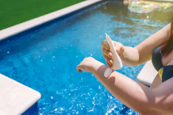 unrecognisable young woman applying sunscreen to the skin on her arms, next to a swimming pool. cosmetic to prevent sunburn. concept of health and skin care. sunlight, outside a garden.