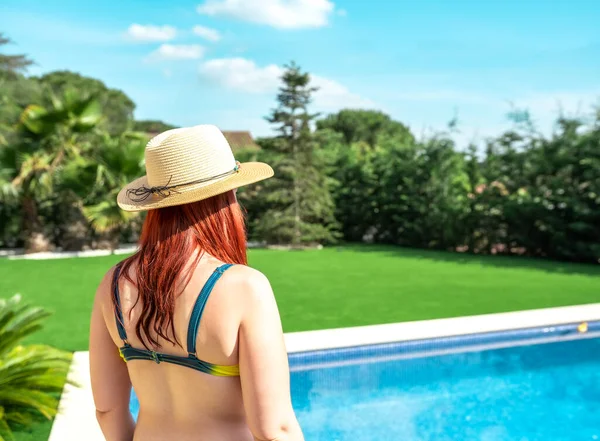 young woman on holiday, with her back to camera looking at the horizon by a swimming pool. girl in summer clothes enjoying her trip. concept of summer and free time. outdoor garden, natural sunlight.