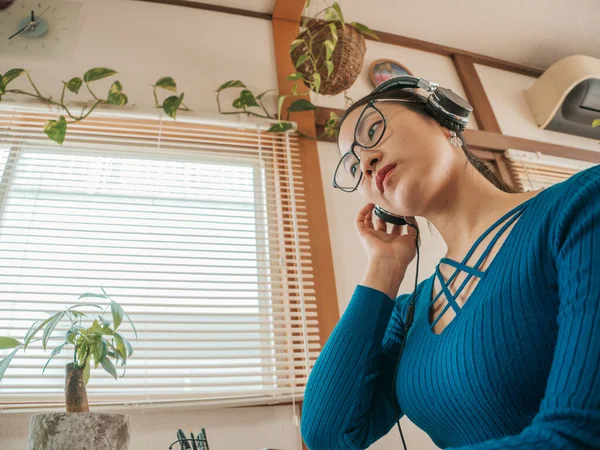 Gözlüklü, kulaklı ve bitkilerli Asyalı kız evde dizüstü bilgisayar ve cep telefonuyla çalışıyor..