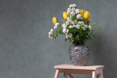 Gri bir vazoda, tahta bir merdivenin gri arka planında bir buket çiçek. Sarı laleler ve beyaz çiçekler. Taze ve renkli çiçekler. Vksna aroması. Evde neşeli bir ruh hali var. Rahatlık ve sükunet.