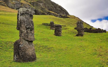 Moai heykelleri, Rano Raraku Volkanı 'nda bulutlu ve yeşil çimenli mavi bir gökyüzüne karşı. Paskalya Adası, Rapa Nui Ulusal Parkı (Şili)