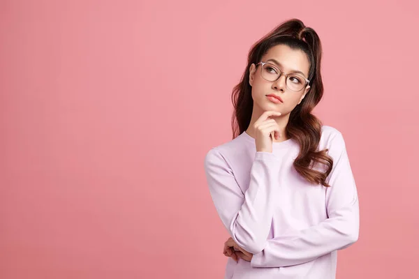 Curious Asian girl is thinking and standing on pink background with curious face, blank copy space for your advertising content.