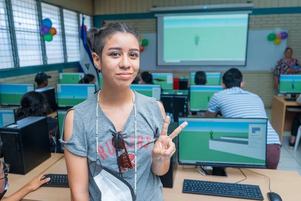 Half-length portrait of a young Latin woman from Central America making a peace symbol with her hand and standing in a classroom full of computers — стокове фото