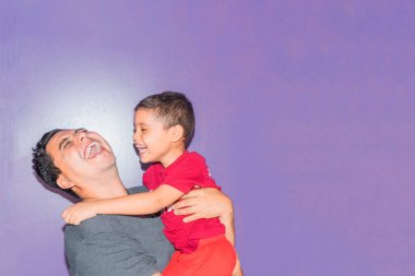 Latin baba gülümsüyor ve oğlunu kollarında mor arka planda taşıyor.