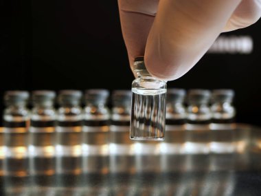 Silikon eldivenli bir el ön taraftaki şişeye dokunuyor, arkasında da bulanık şişeler var. Koyu renkler, sıcak ışık.