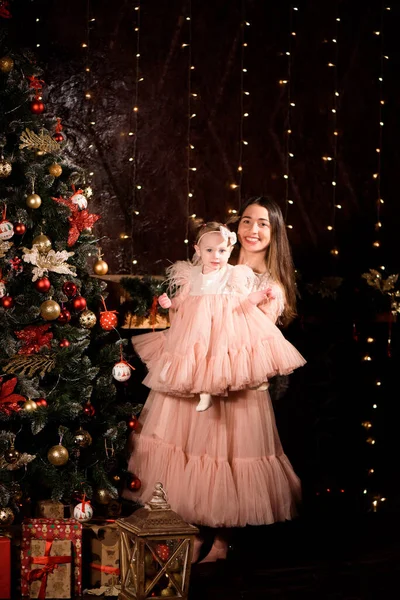 A young mother and daughter in matching dresses decorate the Christmas tree with new balls.