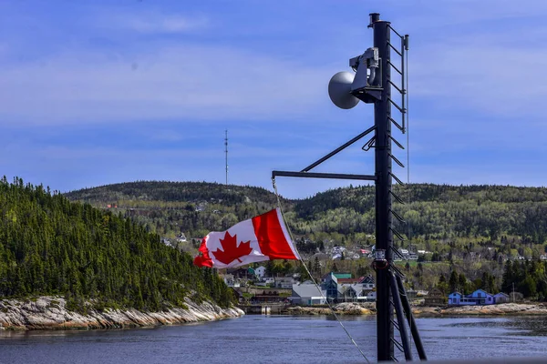 Kanada bayrağına bir feribotla yakın çekim. Arka planda orman ve köy var. Tadoussac, Quebec, Kanada