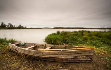 An old wooden boat on the river bank in a remote European location. Brown earth, green grass and cloudy sky. Vintage style photo.