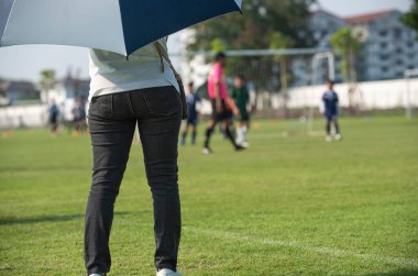 Anne, açık havada ve güneşli bir günde oğlunun okul turnuvasında futbol oynamasını seyrediyor. Spor, aktif yaşam tarzı, mutlu aile ve ev hanımı kavramı..