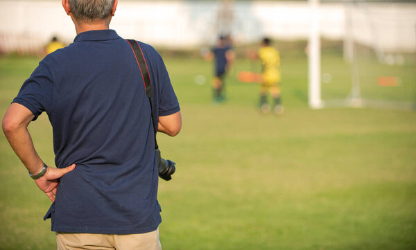 Father standing and watching his son playing football in a school tournament on a clear sky and sunny day. Sport, outdoor active, lifestyle, happy family and soccer mom and soccer dad concept.