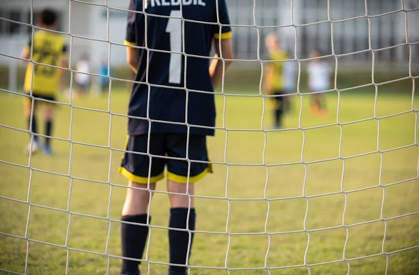 Close up of football goal net, seeing a young soccer goalie goalkeeper during the match. Youth Soccer game on a sunny summer school tournament Day. Football match going on in a background.