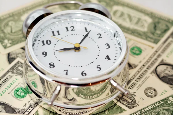An alarm clock on a background of blurred American money banknote currency bills, USA dollars background with a timer in hours, minutes and seconds scale, time is money concept, selective focus