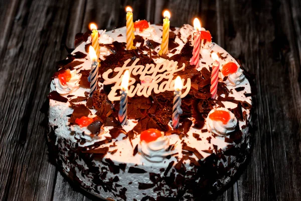 A birthday black forest cake with candles and Happy birthday decoration, chocolate sponge cake, soaked with cherry syrup layered with whipped cream and cherries on a wooden background, selective focus