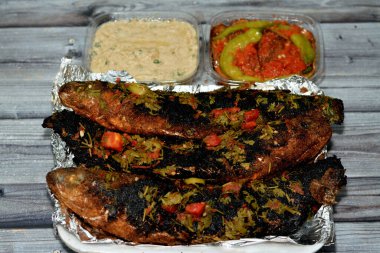 Maydanoz ve domatesli ızgara kefal balığı, patlıcan turşusu ve tahin sosu ızgara ve ahşap arka planda izole edilmiş folyo kağıdında servis edilen tek kullanımlık tabakta.