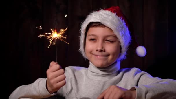 Portrait d'un beau garçon au chapeau rouge avec un scintillement à la main. Enfant avec des lunettes regarde la caméra la veille de Noël. — Video