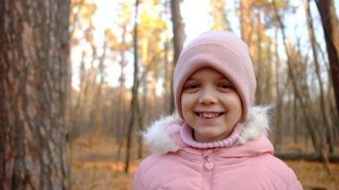 Sonbahar ormanında ceketli ve şapkalı küçük bir kız gülümsüyor. Diş teli takan mutlu çocuk.