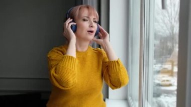 Sarı kazaklı modern kadın kış aylarında evin penceresinde kablosuz kulaklıkla müzik dinler..
