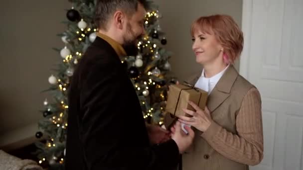 Der Mann schenkt der Frau eine Geschenkbox auf dem Hintergrund des Weihnachtsbaums. Familientradition. — Stockvideo