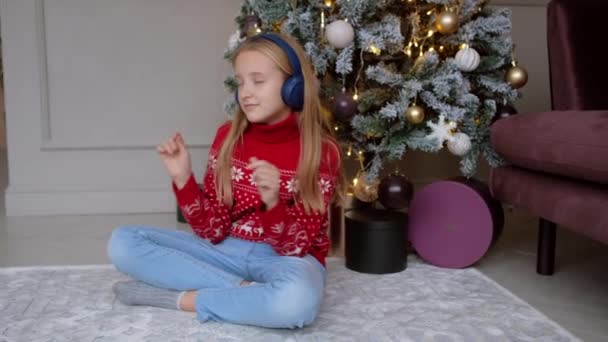 Liten flicka i röd tröja lyssnar på musik eller en ljudbok i trådlösa hörlurar på bakgrunden av julgran. — Stockvideo