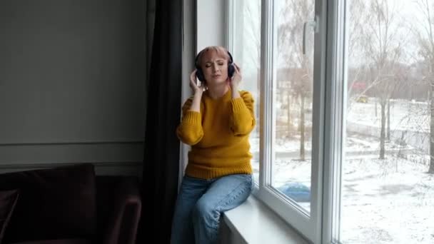 Nowoczesna kobieta w żółtym swetrze słucha muzyki w bezprzewodowych słuchawkach w oknie domu w zimie. — Wideo stockowe