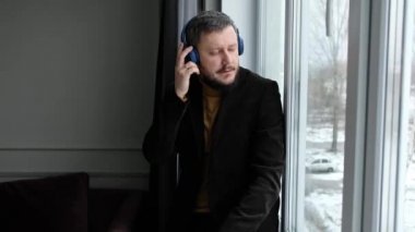 Modern business man in jacket listens to music in wireless headphones at the window of house in winter.