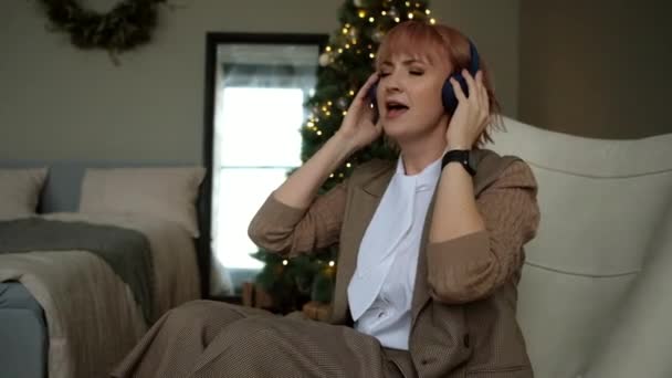Donna d'affari moderna canta e ascolta musica in cuffie senza fili sullo sfondo dell'albero di Natale. — Video Stock