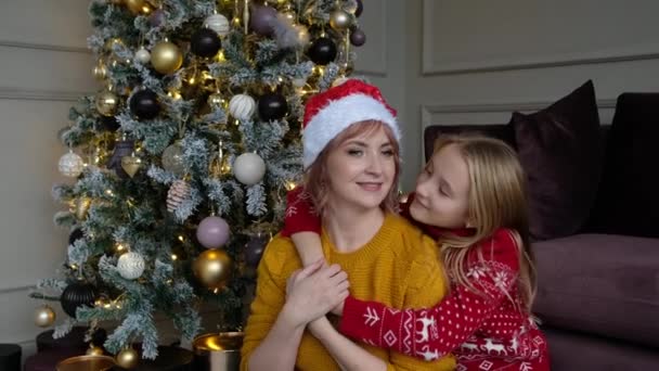 Happy mother and daughter in sweaters and red hat look at camera and smile against background of christmas tree. — Stockvideo