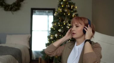 Modern iş kadını Noel ağacının arka planında kablosuz kulaklıkla şarkı söyler ve dinler..