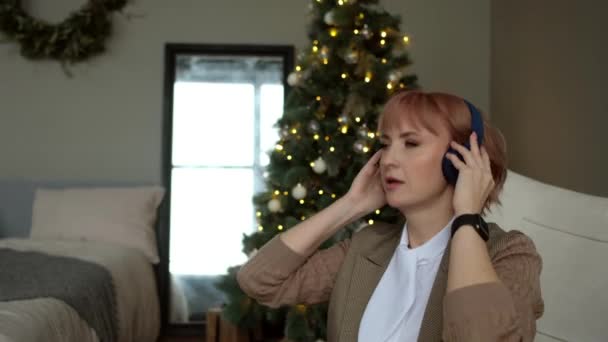 Modern business woman sings and listens to music in wireless headphones on background of Christmas tree. — Video Stock