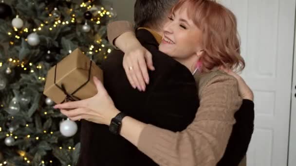 Man gives woman a gift box on background of Christmas tree. Family tradition. — Video
