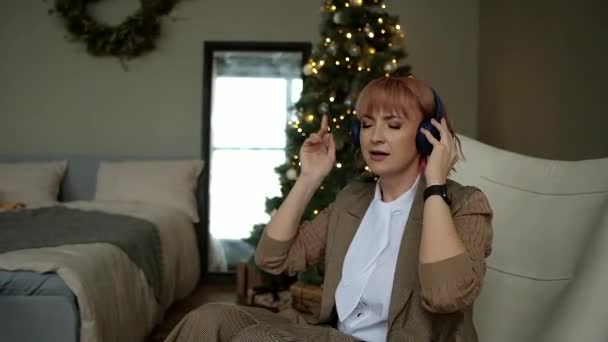 Modern business woman sings and listens to music in wireless headphones on background of Christmas tree. — Αρχείο Βίντεο