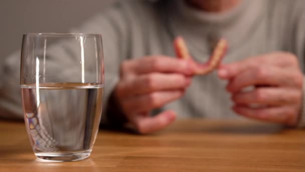 Eine ältere Frau nimmt falsche Zähne aus dem Mund und steckt sie in ein Glas Wasser. Mund-, Prothesen- und Prothesenhygiene. — Stockvideo