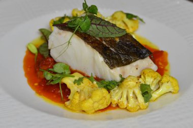 Kırmızı biber soslu morina balığı bifteği ve körili karnabahar, taze otlarla süslenmiş, masadaki bir restoranda servis edilen Akdeniz yemeği.