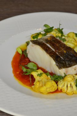 Kırmızı biber soslu morina balığı bifteği ve körili karnabahar, taze otlarla süslenmiş, masadaki bir restoranda servis edilen Akdeniz yemeği.