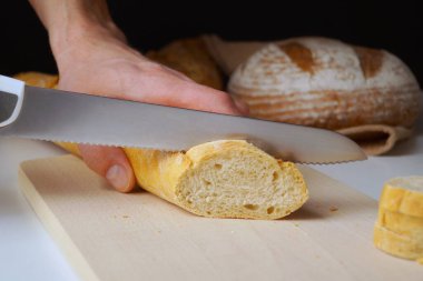 Adamın biri ekmek bıçağıyla baget kesiyor. Arka planda iki somun ekmek daha görülebilir.