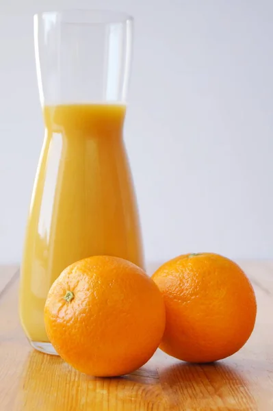 木製のテーブルの上には大きな細長いガラスのジュースが立ち 絞りたてのオレンジジュースが注ぎ込まれています ジャグの隣にはオレンジがあります 一日の始まりと一日のエネルギーとビタミンのブースト — ストック写真