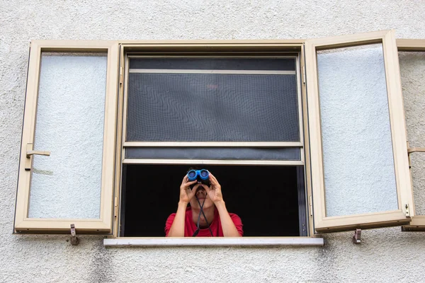 A person with binoculars. A young man investigate or spies on someone with binoculars from the window of a house. A curious person looks at something in the distance