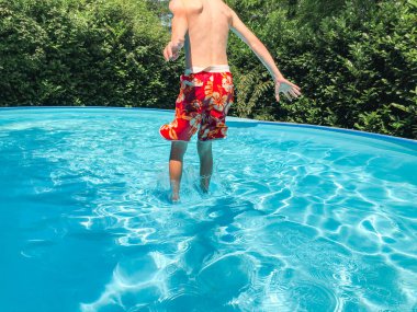 Kırmızı renkli mayo giyen bir çocuk güneşli bir yaz gününde açık mavi şeffaf suyla özel bir havuza atlıyor..