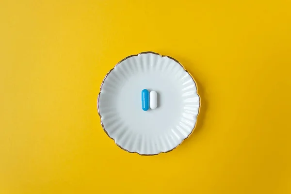 Вид зверху на тарілку з двома білими та синіми капсулами. Жовтий фон і простір для тексту. Добова доза ліків . — стокове фото
