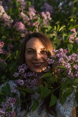 Bahçede mor leylak çiçekleriyle çevrili çekici, gülümseyen beyaz kadın portresi. Bahar çiçeği zamanı. Açık hava moda fotoğrafı. Cottagecore konsepti.