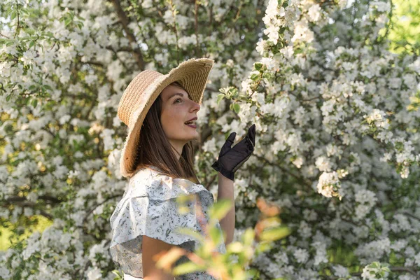 Smiling attractive caucasian woman in retro clothes with straw hat and vintage black lace gloves. Enjoy spring nature, relax in countryside blossom apple tree garden. Cottagecore aesthetics.