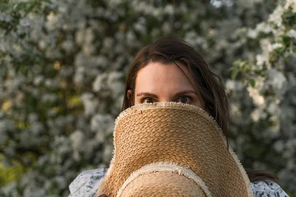 Caucasian woman hiding under a straw hat. Surprised look of brown eyes. Walking in the spring blossom apple tree garden. Cottagecore aesthetics. Fight allergy to seasonal flowering.