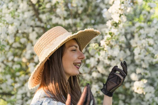 Smiling attractive caucasian woman in retro clothes with straw hat and vintage black lace gloves. Enjoy spring nature, relax in countryside blossom apple tree garden. Cottagecore aesthetics.