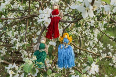 Çiçek açan elma bahçesindeki kırmızı, yeşil ve mavi ipliklerden örülmüş üç el yapımı yüzsüz bebek grubu. Makrame tekniği. Boho tarzında iç dekorasyon. Ukrayna ulusu muskası.