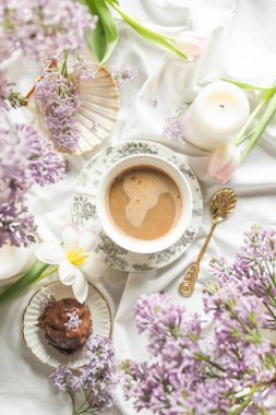 Antik sofra takımı, laleler, leylak çiçekleri, mumlar, deniz kabuğu, bir fincan kahve ve çikolatalı profiterolle yatakta kahvaltı manzarası. Sıcak ev atmosferi. Kadınsı Fransız sabah konsepti.