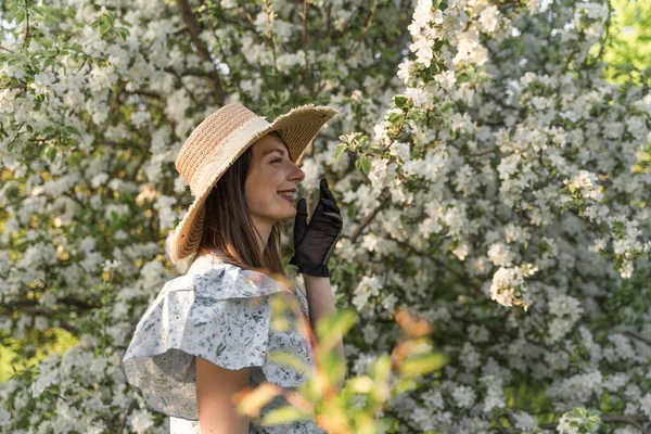 Smiling attractive caucasian woman in retro clothes with straw hat and vintage black lace gloves. Enjoy spring nature, relax in countryside blossom apple tree garden. Cottagecore aesthetics.