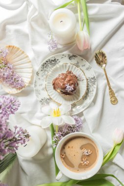 Antik sofra takımı, laleler, leylak çiçekleri, mumlar, deniz kabuğu, bir fincan kahve ve çikolatalı profiterolle yatakta kahvaltı manzarası. Sıcak ev atmosferi. Kadınsı Fransız sabah konsepti.