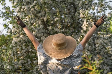 Genç bir kadın, elleri siyah, eski dantel eldivenli hasır şapka tutuyor. Arkadan bak. Bahar çiçeği elma bahçesi. Cottagecore estetiği. Kadın bahar modası kavramı.
