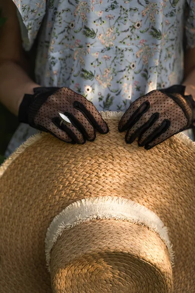 Woman\'s hands in vintage black lace gloves holding a big straw hat. Visual for content. Female spring fashion and boho style. Cottagecore aesthetics concept.