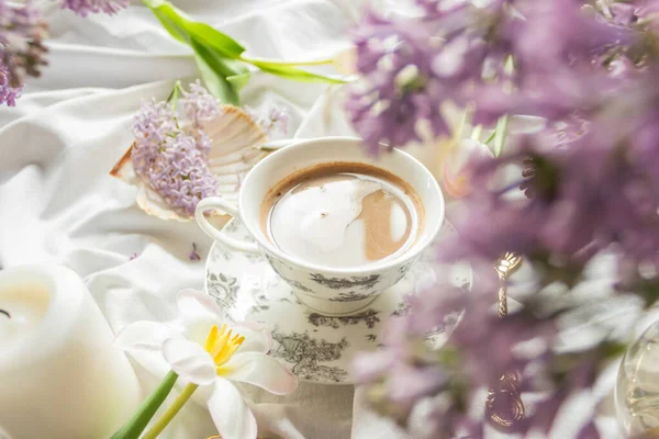 Spring composition with tulips and lilac flowers, candles and cup of coffee on white bed sheets. Cozy home atmosphere. Side view. Feminine morning breakfast concept.
